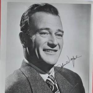 Peut inclure: Photographie en noir et blanc de John Wayne, un acteur célèbre, portant un costume et une cravate. Il sourit et regarde la caméra. Sa signature est écrite à l'encre noire sous son image. Le texte "John Wayne" est imprimé sous la photo.