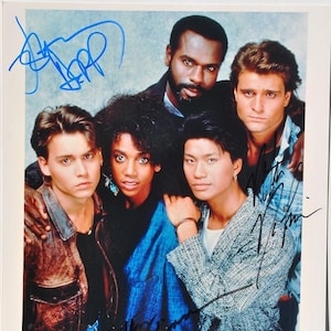 May include: A group of five people, including a man with a beard and a woman with curly hair, are posing for a photo. They are all smiling and looking at the camera. The man in the back is wearing a black shirt and the woman in the front is wearing a blue shirt. The other three people are wearing casual clothing. The photo is signed by all five people.