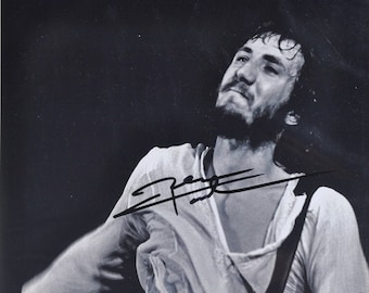 May include: Black and white photo of a musician performing on stage. The musician is wearing a white shirt and has a beard. The musician is playing a black electric guitar and is looking up at the camera. The musician's signature is visible in the top right corner of the image.