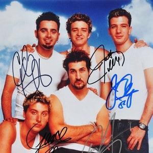 May include: A signed photograph of a boy band with five members. The band members are wearing white shirts and jeans. The photo is signed by each member in blue and black ink.