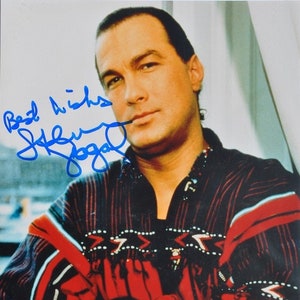 以下が含まれることがあります： 濃い色の髪とあごひげを生やした男性のクローズアップポートレート。彼は幾何学模様の入った赤と黒のストライプのシャツを着ています。男性はカメラにまっすぐ見つめ、真剣な表情をしています。画像は「スティーブン・セガールより、よろしく」と署名されています。