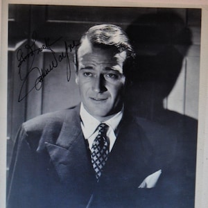 Peut inclure: Photographie en noir et blanc de John Wayne en costume et cravate, avec sa signature en dessous. Le texte indique "John Wayne dans Paramount Pictures".