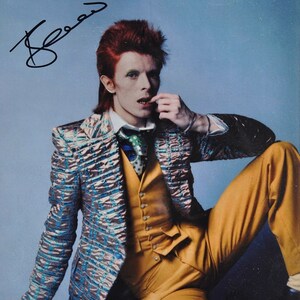 May include: A black and white photo of a person with red hair wearing a blue and gold patterned suit. The person is sitting with their hand near their mouth and has signed their name "Bowie" in the top left corner of the photo.