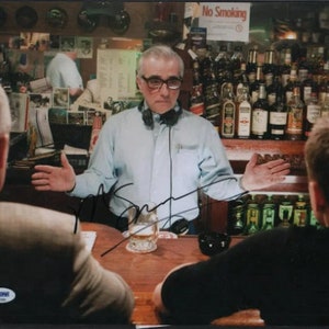 May include: A man wearing a light blue shirt and a microphone is sitting at a bar. He is talking to another man who is wearing a black shirt. There are bottles of alcohol on the shelves behind them. The man in the light blue shirt is signing his name on a piece of paper.