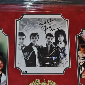 May include: A framed photo of the band Queen with their signatures. The photo shows the band members in black and white, with a red background. The signatures are in blue ink. The frame is black with a gold trim.