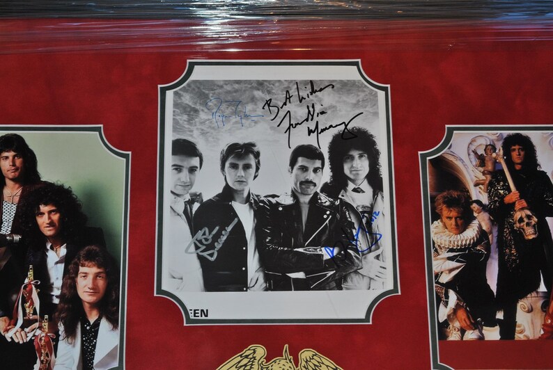 May include: A framed photo of the band Queen with their signatures. The photo shows the band members in black and white, with a red background. The signatures are in blue ink. The frame is black with a gold trim.