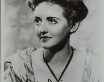 May include: Black and white photo of Bette Davis, a famous actress, with her signature. She is wearing a white blouse and has her hair styled in a bun with flowers.