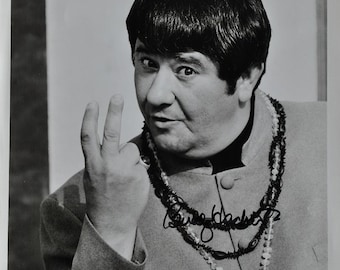 May include: A black and white photo of Buddy Hackett, an American comedian, actor, and musician. He is wearing a dark colored shirt with a beaded necklace and is making a peace sign with his fingers. The photo is from the 1968 film "The Love Bug".