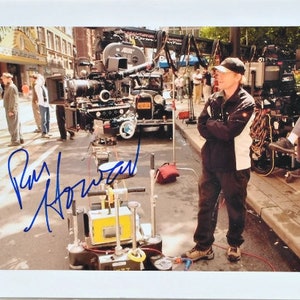 May include: A black and white photograph of a film set with a director standing in the middle of the street. The director is wearing a black jacket, a white shirt, and a black baseball cap. The director is holding his arms crossed and looking to the right. The director's name is written in blue ink on the bottom left corner of the photo: Ron Howard.
