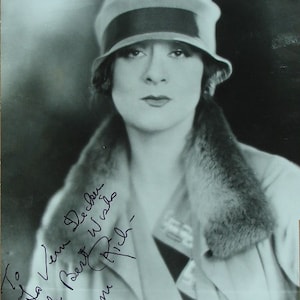 May include: Black and white portrait of a woman wearing a black and white hat with a fur collar. The woman is looking at the camera and has a neutral expression. The image is signed "To Ida Ven Decher with Best Wishes,  Gena Rich."