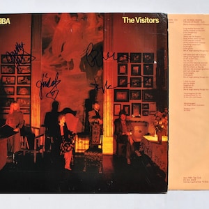Könnte beinhalten: Das Albumcover von ABBA's "The Visitors" zeigt eine Gruppe von Personen in einem Raum mit Unterschriften. Das Cover hat ein dunkles Rot- und Braunschema. Der Albumtitel ist in weißer Schrift. Ein Textblatt befindet sich daneben.