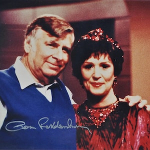 May include: A black and white photo of a man and a woman. The man is wearing a blue sweater vest and a white shirt. The woman is wearing a red dress with a jeweled necklace. The man is signing his name "Ben  Robertson" in blue ink.
