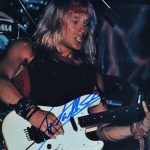 May include: A live performance photo of a musician playing a white electric guitar. The musician is wearing a black tank top, a red bandana, and wristbands. The guitar has a black tremolo system and is signed in blue ink.