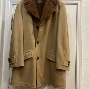 Peut inclure: Un manteau en laine beige avec un col en fausse fourrure marron. Le manteau a une fermeture à trois boutons, deux poches avant et des poignets boutonnés. Il est suspendu à un cintre métallique contre une porte blanche.