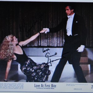 Könnte beinhalten: Ein Schwarzweißfoto von einem tanzenden Paar. Die Frau trägt ein schwarzes Kleid mit weißem Muster und der Mann trägt einen schwarzen Smoking. Das Foto ist signiert mit "Love Gene Simmons". Der Filmtitel "Love At First Bite" ist unten auf dem Foto gedruckt.