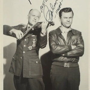 May include: Black and white photo of two men, one playing a violin and the other with his arms crossed. The man playing the violin is wearing a military uniform with medals. The man with his arms crossed is wearing a leather jacket. The photo is signed "Best Writers" and "Phil Silvers - Hope."