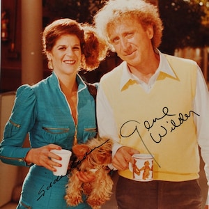 May include: A color photograph of a man and a woman standing next to each other. The man is wearing a yellow sweater vest over a white shirt and brown pants. The woman is wearing a blue jumpsuit and holding a small dog. The man is holding a mug with a picture of two people on it. The man has signed his name "Gene Wilder" and the woman has signed her name "Gilda Radner".