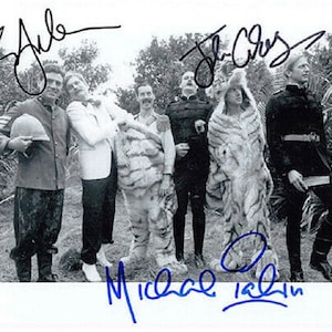 Op de afbeelding: Zwart-witfoto van vijf mannen in een jungle-omgeving. De mannen dragen verschillende outfits, waaronder een wit pak, een tijgerkostuum en militaire uniformen. De foto is gesigneerd door de mannen, waaronder Michael Palin, John Cleese en Terry Gilliam.