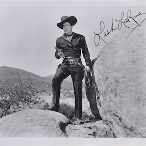 May include: Black and white photo of a man wearing a cowboy hat and holding a gun. He is standing on a rock with a large rock behind him. The photo is signed "Lash LaRue".