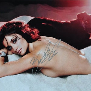 Pode incluir: Fotografia de uma mulher deitada numa superfície arenosa, com um cão de cor escura atrás dela. A mulher tem cabelo escuro e usa uma pulseira. A imagem tem um autógrafo no canto inferior direito.