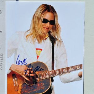 Può includere: Una donna con occhiali da sole e una giacca bianca suona una chitarra acustica marrone. La chitarra ha un battipenna nero e un motivo floreale. La firma "Love Avril" è scritta in inchiostro blu sulla chitarra.