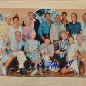 May include: A group of people, likely a cast or crew, pose for a photograph. The image features a variety of ages and attire, with some individuals wearing casual shirts and others in more formal wear. The photo is signed in blue ink.