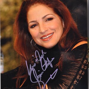 May include: A woman with long, auburn hair smiles at the camera. She is wearing a black, lace-trimmed top. Her autograph is written in purple ink below her image.