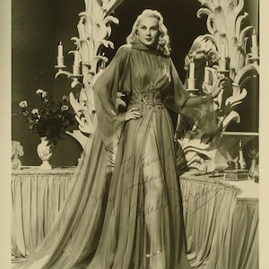 Puede incluir: Una fotografía en blanco y negro de una mujer con un vestido largo y fluido con la cintura ajustada. Está de pie frente a un telón de fondo decorativo con velas y detalles ornamentados. La mujer sonríe y mira a la cámara.