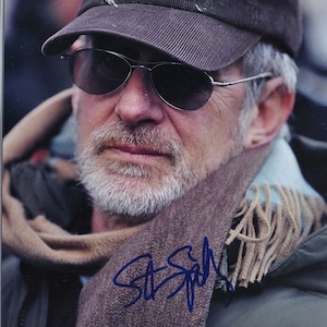 May include: A man wearing sunglasses, a brown corduroy baseball cap with "UNIVERSITY" text, and a scarf. The scarf is brown with blue fringe. The image includes a signature in blue ink.