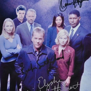 May include: A group of seven people, including actors from the TV show 24, pose for a photo. The actors are standing in a line, with the exception of the person in the center, who is standing in front of the others. The actors are all smiling and looking at the camera. The photo is signed by all of the actors.