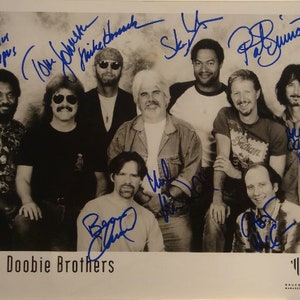 May include: A black and white photo of The Doobie Brothers band members. The band members are all smiling and looking at the camera. The photo is signed by all of the band members. The band members' names are listed below the photo:  "The Doobie Brothers".