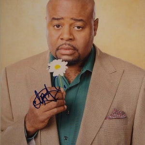 May include: A man in a tan suit jacket with a green shirt and a white flower in his hand. He is smiling and looking at the camera. The image is signed with a blue signature.