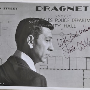 May include: Black and white photograph of Jack Webb, the star of the television show Dragnet, with a handwritten inscription "With Best Wishes Jack Webb". The photo is set against a backdrop of a building layout.