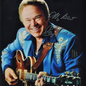 Puede incluir: Un hombre con una chaqueta azul con bordados plateados toca una guitarra eléctrica marrón y dorada. Sonríe y ha firmado su nombre "Mr. Best" con tinta plateada.