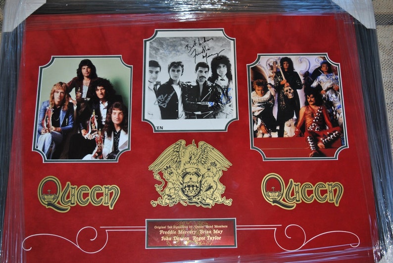 May include: A framed red background with three black and white photos of the band Queen. The photos are surrounded by gold trim. The band's crest is in the center of the frame. The text "Queen" is in gold lettering at the bottom of the frame. The text "Original Ink Signatures by Queen Band Members Freddie Mercury Brian May John Deacon Roger Taylor" is at the bottom of the frame.
