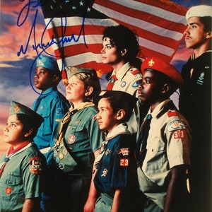 May include: A group of children wearing scout uniforms stand in front of an American flag. The children are all smiling and looking at the camera. The flag is waving in the wind. The image is signed by Michael Jackson.