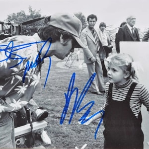 Könnte beinhalten: Ein Schwarzweißfoto eines Mannes mit Baseballkappe und Blumenhemd, der einer jungen Frau mit gestreiftem Shirt und Latzhose ein Autogramm gibt. Der Mann schaut auf das Mädchen, und das Mädchen schaut zu dem Mann auf. Im Hintergrund sind weitere Personen zu sehen.