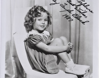 May include: Black and white photograph of Shirley Temple, seated on a decorative chair. The image includes an autograph that reads "To Mike Lefebvre, Best Wishes, Shirley Temple Black, 1987." The chair has a star design and fringe.
