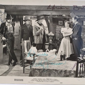 May include: Black and white photograph of a scene from the film "The Searchers". The image features four actors, including John Wayne, in a dimly lit room. The actors are standing around a table with a pitcher and glasses. The image is signed by the actors, including John Wayne, Jeffrey Hunter, and Ward Bond.
