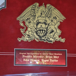 May include: A red background with a gold Queen logo featuring a phoenix and two lions. The logo is surrounded by a gold frame. Below the logo is a gold plaque with the text "Original Ink Signatures by "Queen" Band Members Freddie Mercury Brian May John Deacon Roger Taylor".