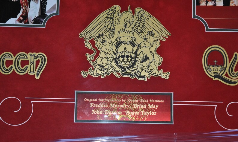 May include: A red background with a gold Queen logo featuring a phoenix and two lions. The logo is surrounded by a gold frame. Below the logo is a gold plaque with the text "Original Ink Signatures by "Queen" Band Members Freddie Mercury Brian May John Deacon Roger Taylor".