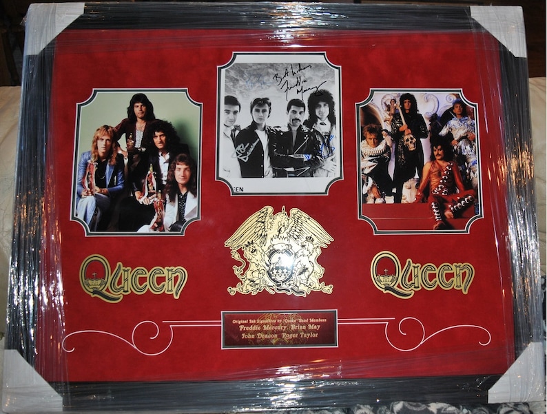 May include: A framed Queen band memorabilia featuring a red background with gold accents. The frame includes three photos of the band members, a gold Queen crest, and the band's name in gold lettering. The photos include a group shot of the band, a close-up of the band members' signatures, and a photo of the band performing on stage. The text below the photos reads "Original UK Exclusive Band Members Freddie Mercury, Brian May, John Deacon, Roger Taylor."