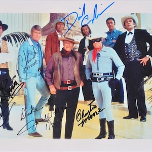 Peut inclure: Un groupe de sept hommes en tenue de cow-boy, dont un chapeau de cowboy et un masque, se tiennent ensemble dans une pièce avec un palmier en arrière-plan. Les hommes sourient tous et regardent la caméra. L'image est signée par les sept hommes, dont "Chuck Connors", "Clint Walker", "Don Collier", "James Drury", "Robert Fuller", "David Hedison", et "Blackie Moore".