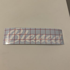 May include: White vinyl decal with the word "Blessed" in a cursive font on a blue gridded transfer sheet.