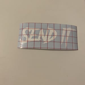 May include: White vinyl sticker with the words "SEND IT" in a bold, stylized font. The sticker is on a gridded backing.