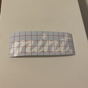 Può includere: Adesivo in vinile bianco con la parola "mint" scritta in corsivo su uno sfondo trasparente con un motivo a griglia rossa.