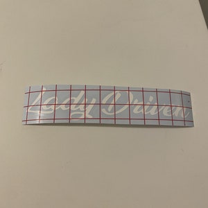 May include: White vinyl sticker with the words "Lady Driven" in a cursive font on a blue background with red grid lines.