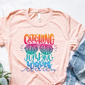 May include: A peach-colored t-shirt with the text "Catching Rays and Jumping Waves" in rainbow colors. The text is stylized with a pair of sunglasses and a wavy line underneath.