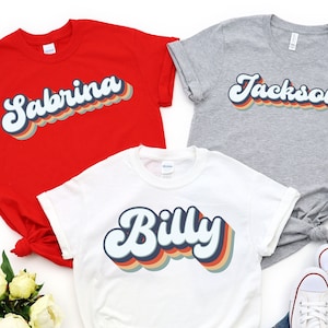 May include: Three t-shirts with retro-style names printed on them. The shirts are red, white, and grey. The names are Sabrina, Billy, and Jackson.
