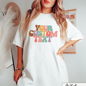 Op de afbeelding: Wit oversized T-shirt met de tekst "YOUR CUSTOM TEXT" in kleurrijke, retro-stijl letters.
